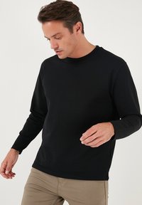 T-shirt nero a maniche lunghe in cotone, con collo rotondo e design aderente, indossato con pantaloni chiari. Texture liscia, senza pattern.