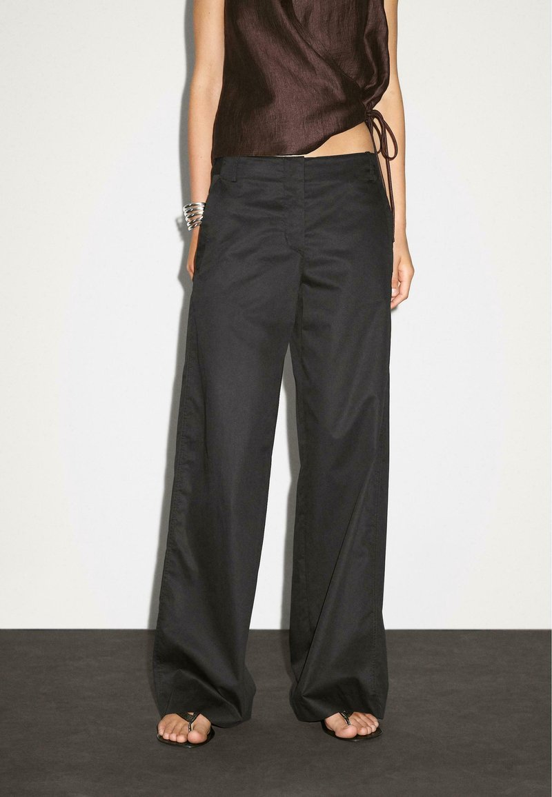 Personne portant un pantalon large noir ample et un haut sans manches marron foncé avec des sandales sur un sol sombre et un fond clair.