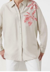 Chemise boutonnée en tissu beige clair ; présente un imprimé floral rouge sur l'épaule gauche, des manches longues et une coupe décontractée.