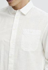Camisa blanca de botones, hecha de una tela ligera y texturizada. Presenta un cuello clásico, un bolsillo en la parte delantera y un diseño sutil y transpirable.