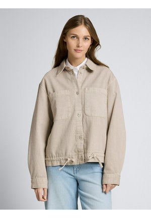 Junge Frau mit langen Haaren, die eine beige Jacke mit Knöpfen über einem weißen Hemd und hellblauen Jeans trägt, steht vor einem schlichten Hintergrund.