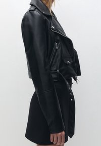 Černá kožená motorkářská bunda s výraznými zipy, zatrhávacími knoflíky, širokým límcem a texturovanými rukávy, doplněná jednoduchou černou sukní.