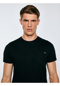 Noor mees, lühikeste pruunide juustega, seljas musta värvi kitsas T-särk rinnataskuga, vaatab otse kaamerasse heleda tausta ees.