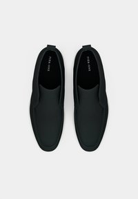 Chaussures slip-on noires avec une texture lisse et des détails de couture. La semelle est blanche, contrastant avec la partie sup érieure. Le nom de la marque est visible à l'intérieur.