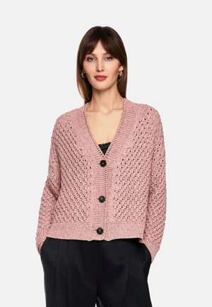 Woman wearing a pink knitted buttoned cardigan with a V-neck and black pants, standing with hands in pockets against a white background.