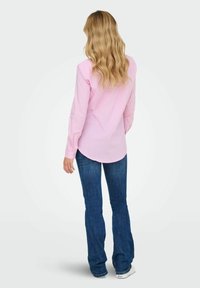 Camisa de rayas rosa de manga larga con puños abotonados, combinada con jeans de mezclilla oscuros acampanados y zapatillas blancas, vista desde la parte trasera.