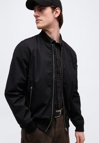 Jeune homme portant une casquette noire et une veste zippée noire sur une chemise à carreaux, avec un pantalon marron et une ceinture à boucle argentée, regardant vers la gauche.