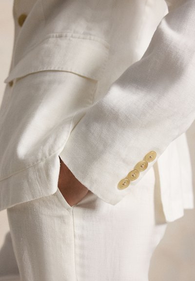 Χέρι σε λευκό σακάκι από λινό με τέσσερα μπεζ κουμπιά στο μανίκι, το χέρι ακουμπάει στην τσέπη του αντίστοιχου λευκού λινού παντελονιού.