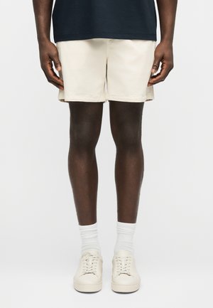 Man wearing beige shorts, white socks, and beige sneakers, standing against a plain light background.