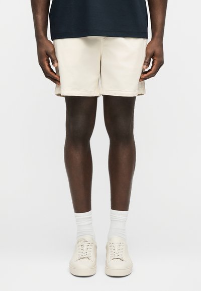 Homme portant un short beige, des chaussettes blanches et des baskets beiges, debout devant un fond clair uni.
