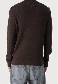 Hnědý pletený svetr s vysokým límcem, který má strukturovaný vzor a lemy na rukávech. Nosit s bledě modrými, volnými džíny.