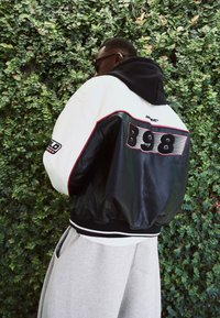 Giubbotto bomber in pelle nera e tessuto crema con accenti rossi, caratterizzato da una grande grafica "898" e polsini a coste. Indossato sopra pantaloni felpati grigi.