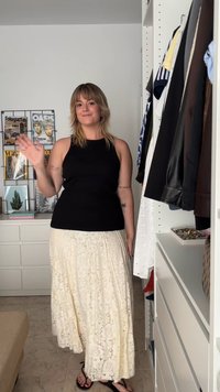 Femme aux cheveux mi-longs portant un haut sans manches noir et une jupe en dentelle crème, faisant signe, debout à côté d'une armoire et d'une commode blanches.