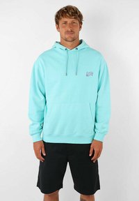 Haut à capuche bleu clair avec une poche avant, un cordon de serrage sur la capuche et un petit logo sur la poitrine. Accompagné de shorts noirs.