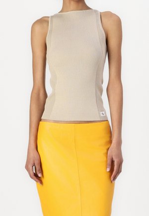 Kobieta ubrana w bezrękawnikowy beżowy prążkowany top i jasną żółtą spódnicę z wysokim stanem, stojąca na tle jednolitego białego tła.