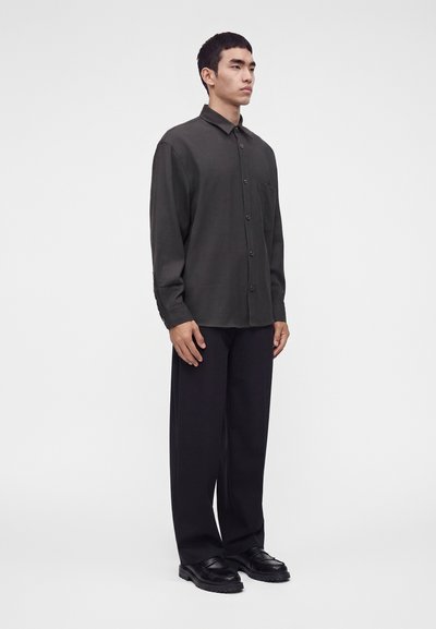 Homme debout portant une chemise sombre à manches longues boutonnée, un pantalon noir ample et des chaussures noires sur un fond clair uni.