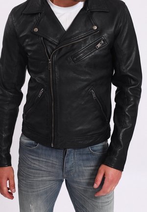 Homme portant une veste de motard en cuir noir avec des poches zippées sur une chemise blanche et un jean bleu délavé.