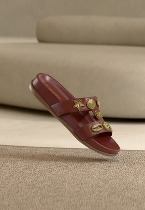 Ruda šlepetė su dviem dirželiais, puošta auksinėmis jūros kriauklių ir žvaigždžių žavėmis, šiek tiek pakibusi virš šviesiai rudos spalvos kilimo.