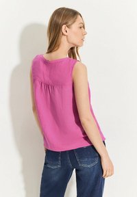 Rosa ärmlös topp med rund halsringning och rynkdetalj på baksidan, matchad med mörkblå jeans med bakfickor.