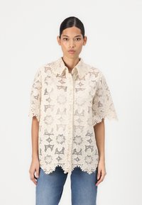 Camicia in pizzo color beige chiaro con motivi floreali, maniche corte e bordi ondulati. Indossata sopra pantaloni di denim blu.