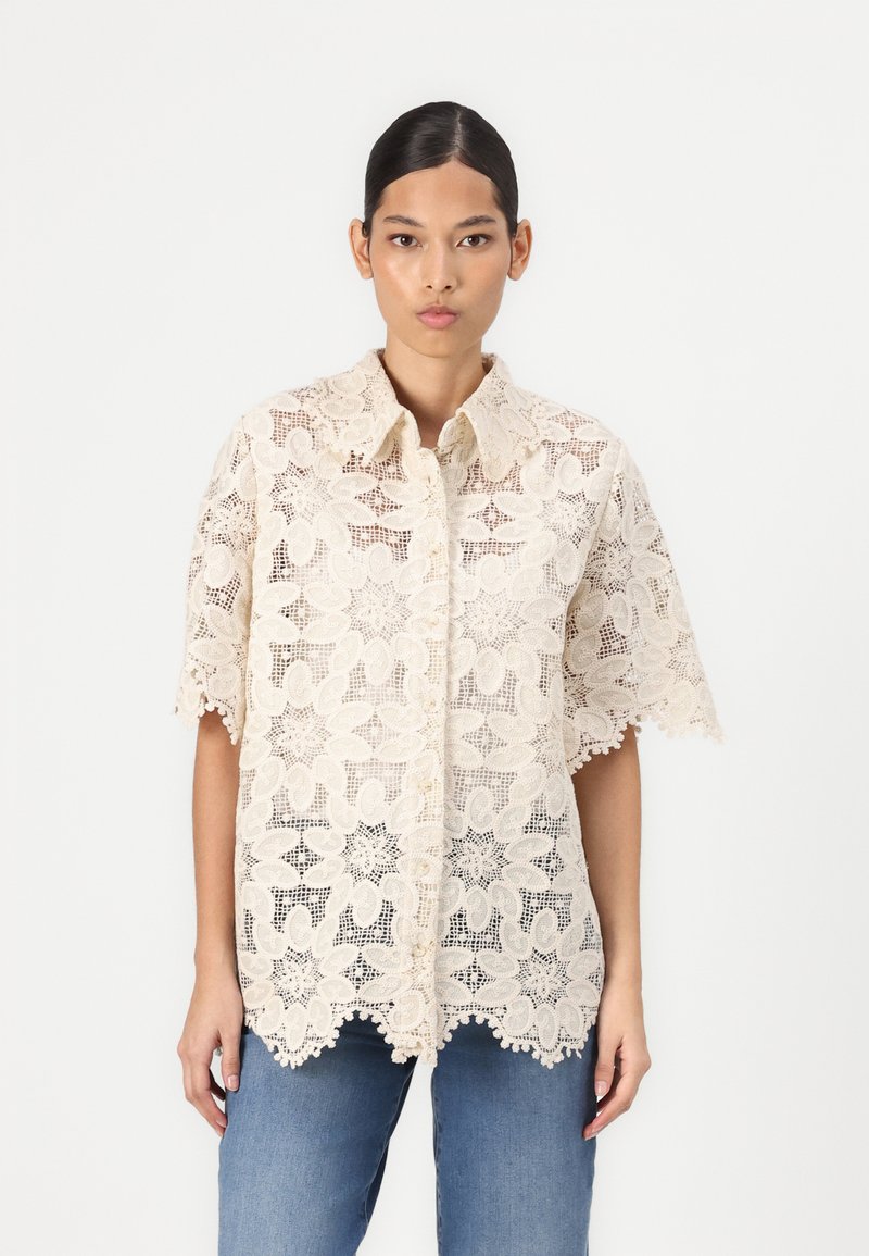 Camicia in pizzo color beige chiaro con motivi floreali, maniche corte e bordi ondulati. Indossata sopra pantaloni di denim blu.