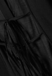 Vêtement noir en tissu texturé avec des franges longues et effilochées le long du côté, créant un effet visuel en couches.