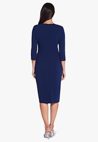 Tummansininen mekko, jossa on kolme neljänneshihaa, istuva vyötärö, polvenpituinen siluetti ja takana vetoketj yksityiskohta. Yhdistetty hopeisiin korkokenkiin.