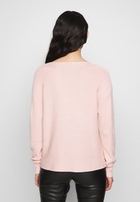 Femme aux longs cheveux noirs portant un pull en maille rose clair et un pantalon en cuir noir, vue de dos sur un fond uni.