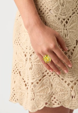 Yellow octagonal ring with a faceted surface worn on a hand, set against a beige crochet lace background with floral patterns.