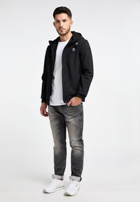 Černá kapucí bunda se zapínáním na zip v přední části a bočními kapsami, v kombinaci se světle šedými džíny a bílými teniskami. Minimalistický design a hladká textura.
