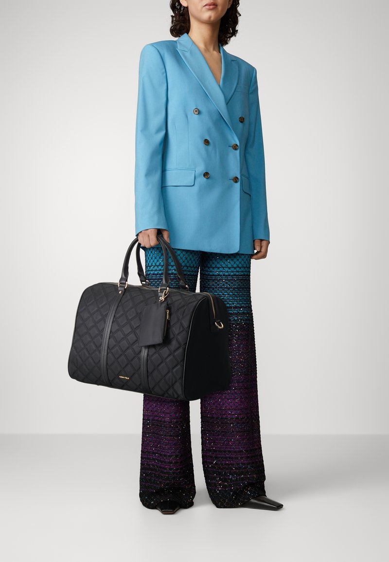 Osoba v modrém dvouřadém saku a širokých kalhotách s gradientem modré a fialové, držící velkou černou prošívanou kabelku.