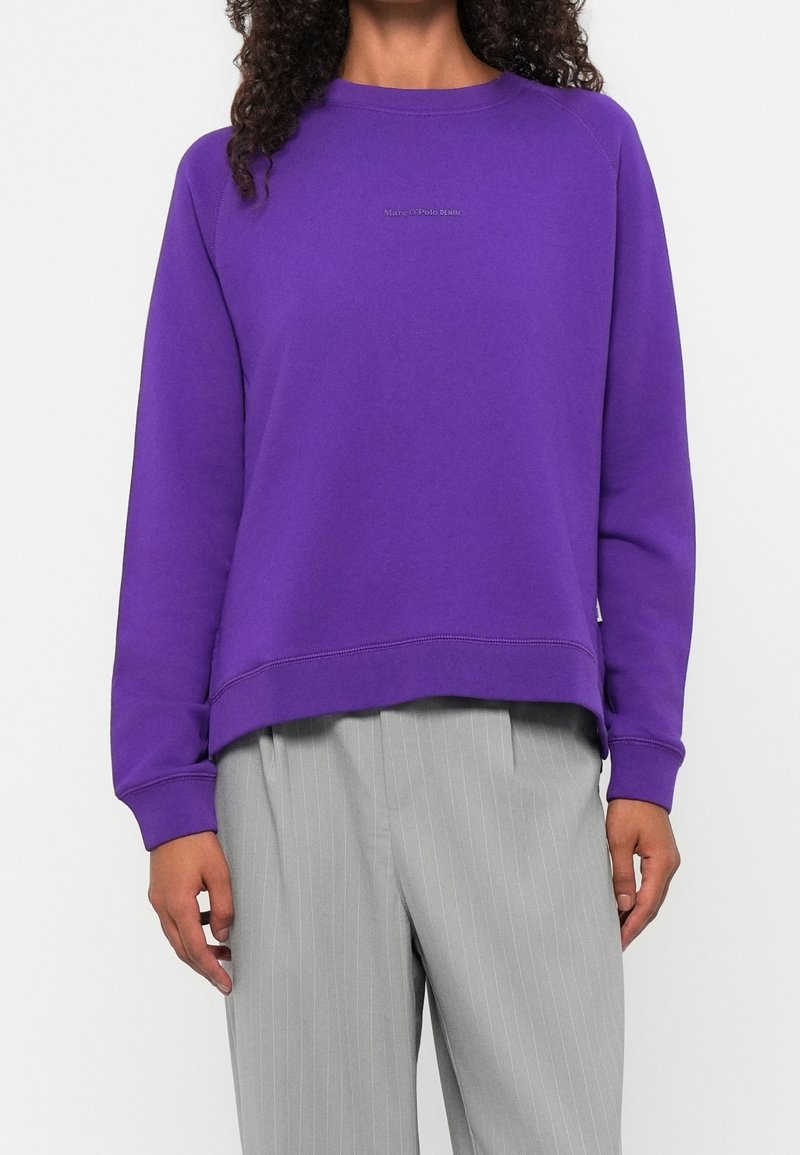 Personne portant un sweat-shirt violet uni à manches longues et un pantalon gris clair à fines rayures, se tenant devant un fond blanc.