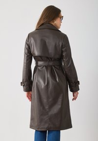 Brown leather trench coat with a belted waist, notched collar, and long sleeves. Smooth texture with pleats at the back.