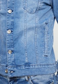 Close-up van een blauwe denim jasje met zilveren knopen en zichtbare stiksels, gedragen over bijpassende blauwe jeans.