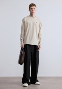 Bézs hosszú ujjú ing apró fekete logóval, laza fekete nadrággal és fehér tornacipővel párosítva. A modell egy barna táskát tart.