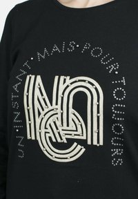 Camisa de algodón negra con un gran gráfico blanco estilizado que presenta curvas y líneas, decorada con tachuelas plateadas y texto impreso.
