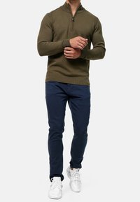 Maglione zip-up verde oliva con polsini e orlo a costine, abbinato a pantaloni slim-fit blu navy e sneakers bianche, creando un look casual.