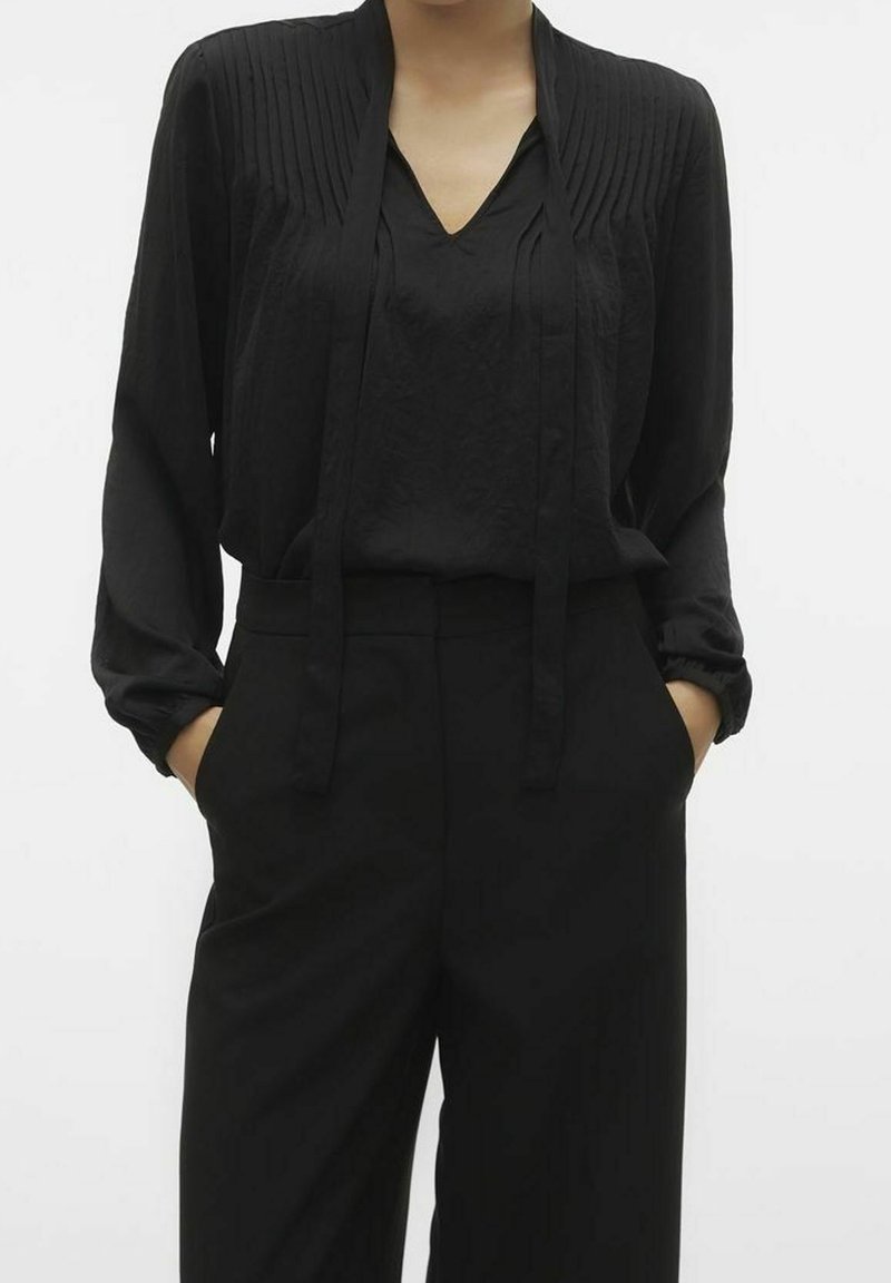 Blouse noire avec plis verticaux, col en V profond et manches longues. Associée à un pantalon noir taille haute, tous deux en tissu lisse.