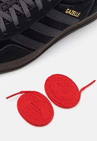 Czarny zamszowy sneaker Adidas Gazelle z czarnymi skórzanymi paskami i złotym logo; obok buta znajdują się czerwone, okrągłe sznurówki.