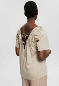 Personne aux cheveux courts portant un haut beige à manches courtes avec un dos ouvert orné de deux nœuds noués et un pantalon beige ample.