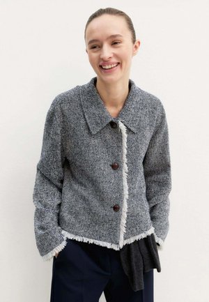 Donna sorridente che indossa una giacca grigia con texture, tre bottoni e bordi sfrangiati, in piedi con le mani nelle tasche davanti a uno sfondo semplice.