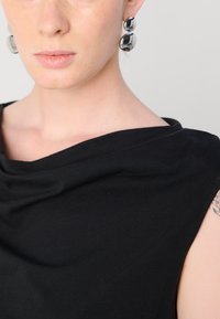 Black, draped top with a wide neckline. Smooth fabric, sleeveless design. Silver circular earrings complement the look.