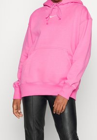 Rosa huvtröja med dragsko, framficka och vit logotyp. Har ribbade muddar och nederkant, tillverkad av mjukt material, med lös passform.