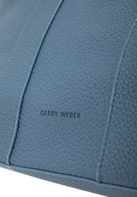 Acercamiento de un panel de bolsa de cuero azul texturizado con costuras verticales y el nombre de la marca "Gerry Weber" grabado en el centro.