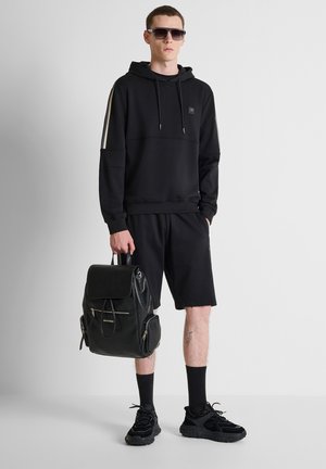 Mann trägt schwarzen Hoodie, Shorts und Turnschuhe, hält einen schwarzen Lederrucksack und steht vor einem schlichten hellen Hintergrund mit Sonnenbrille.