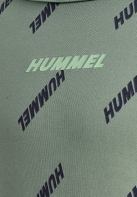 Grüner Stoff mit einer gerippten Textur, der wiederholt im Dunkelblau bedruckte "HUMMEL"-Logos zeigt, die diagonal über die Oberfläche angeordnet sind.