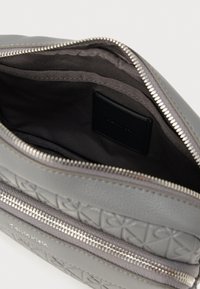 Grå tekstureret lædertaske med lynlåslukning, der har en glat inderside og en sort labeldetalje. Designet med en slank, minimalistisk form.