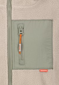 Section de veste en polaire gris clair avec poche zippée verticale dotée d'une tirette orange et d'une étiquette de marque "SUBLEVEL" rouge.