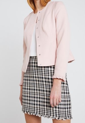 Vrouw draagt een lichtroze jas met knopen over een wit shirt en een zwart-witte geruite rok met een franjes zoom.