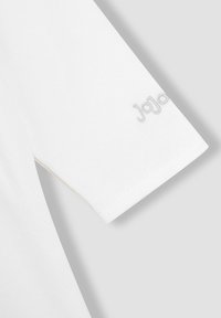 T-shirt blanc en coton avec des manches courtes ; présente un logo "JoJo" brodé gris subtil sur la manche. Texture lisse et design épuré.